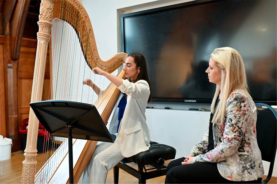 A female student, wearing a white suit, playing the harp, with a women with blonde hair, sitting next to her observing the student.